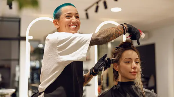A woman with short blue hair applies hair dye and foil to the hair of a woman sitting in a salon chair