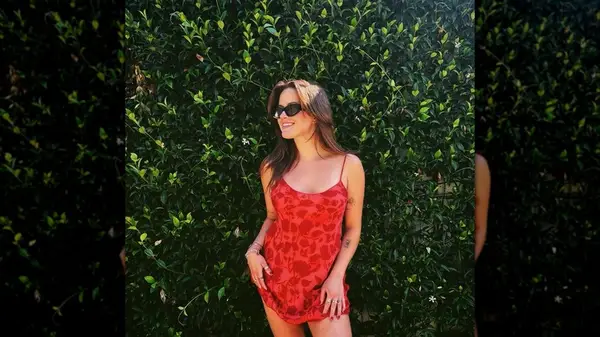 Ava Phillippee smiling in front of a tall hedge in an orange dress that shows some of her arm tattoos