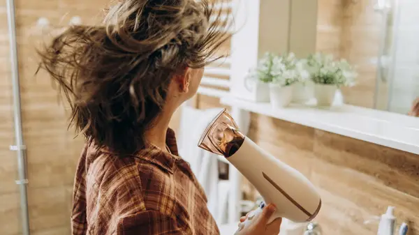 A woman blowdrying her brown hair in a bright bathroom