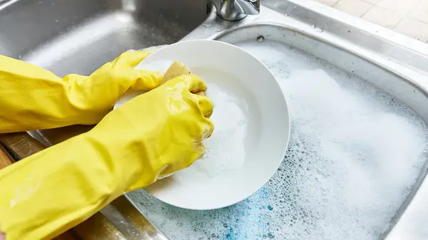 Close-up of hands wearing yellow gloves as they wash a plate over a sink full of soapy water