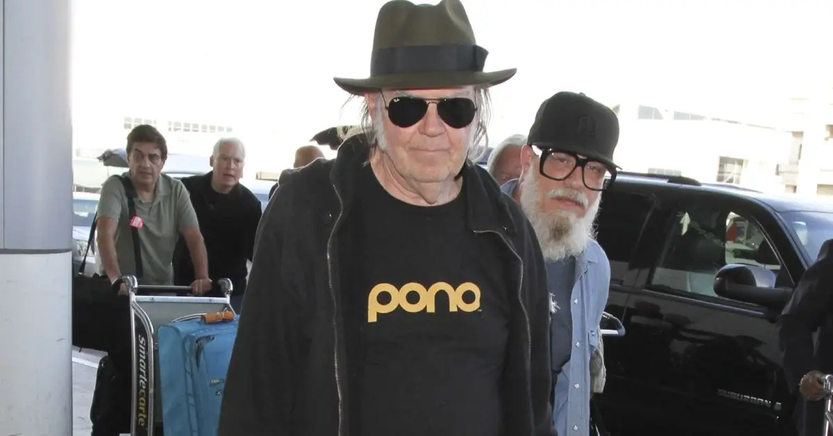 Neil Young trägt Sonnenbrille und Hut vor einem Flughafen