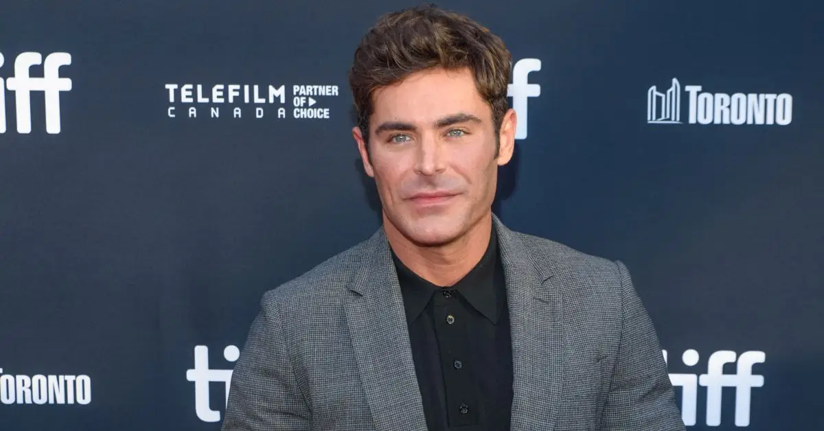 Zac Efron vestindo camisa preta e jaqueta cinza