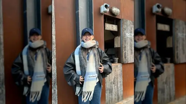 A woman wearing a ball cap, leather jacket and oversized scarf standing in front of wall