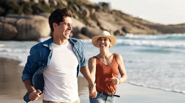 A couple running on the beach holding hands while the woman wears a hat