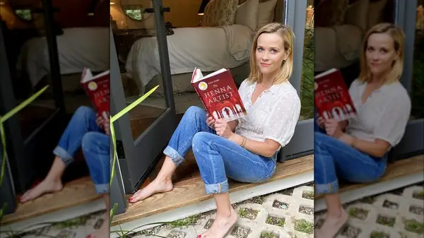 Reese Witherspoon sitting by an open door in jeans and a white top while holding a book