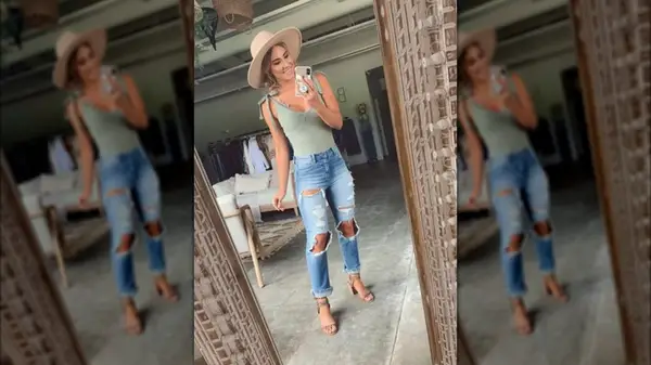 A woman photographs herself in the mirror wearing a hat and distressed jeans