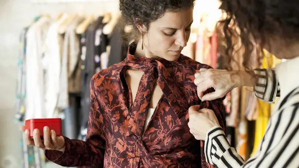 A woman looks down as another woman stitches her dress at the shoulder