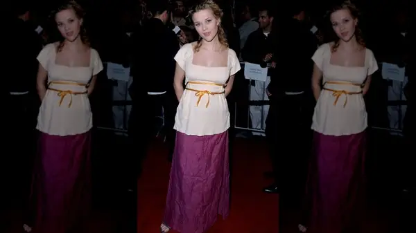 Reese Witherspoon smiling with her hand behind her back, wearing a bloude and purple skirt