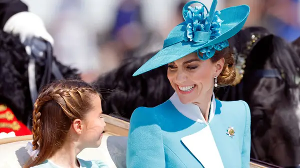 Kate Middleton, in a blue suit and hat, smiles down at Charlotte while both sit in a parade coach