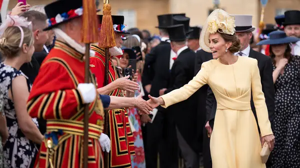 Kate Middleton wearing a yellow dress while reaching to grasp a fan