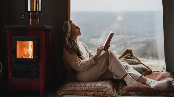 Cozy-looking woman reading fireside in front of a window