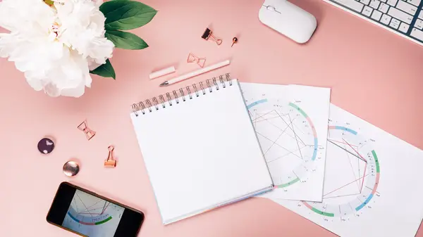 Birth charts laid out on a pink table with a phone and notebook