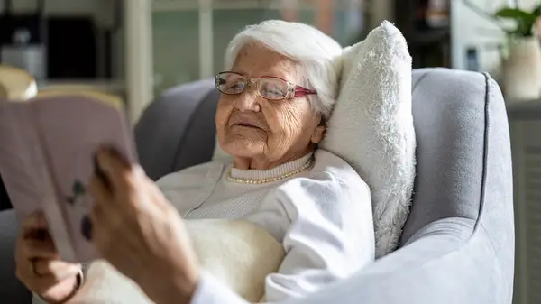 An elderly woman rests comfortably on a chair as she reads a book