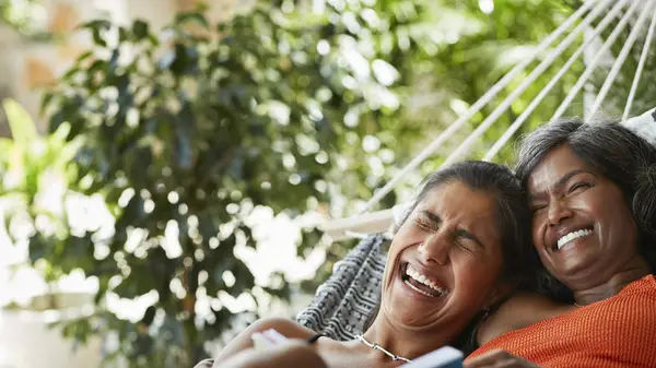 Two women share a hammock and laugh while holding a book