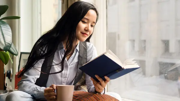 A young Asian woman sitting in the window and reading a book while holding a mug