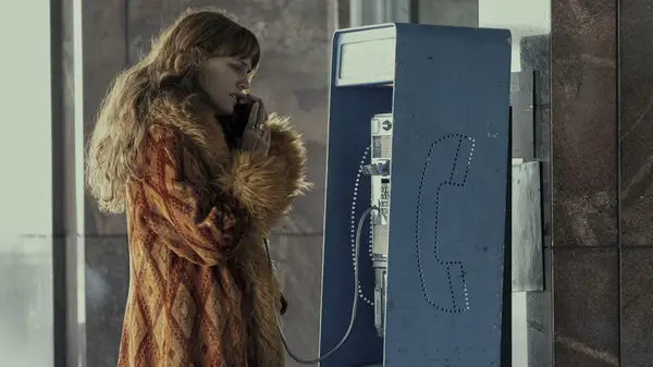 A woman in an oversized fur coat talking to a pay phone