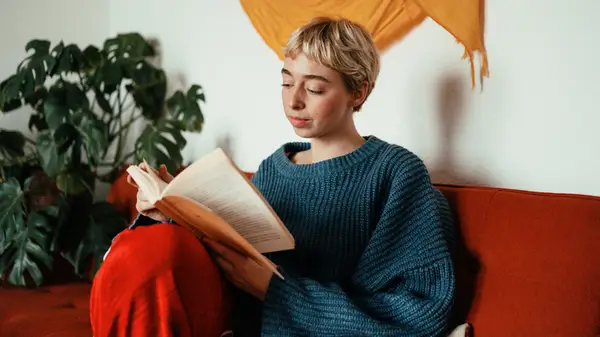 A blonde woman sitting on a red couch and reading a book
