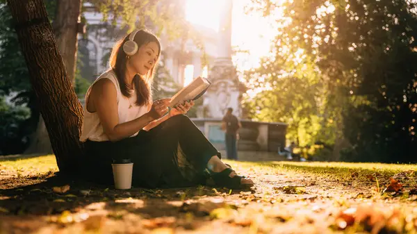 A young woman enjoys a book with headphones on outside