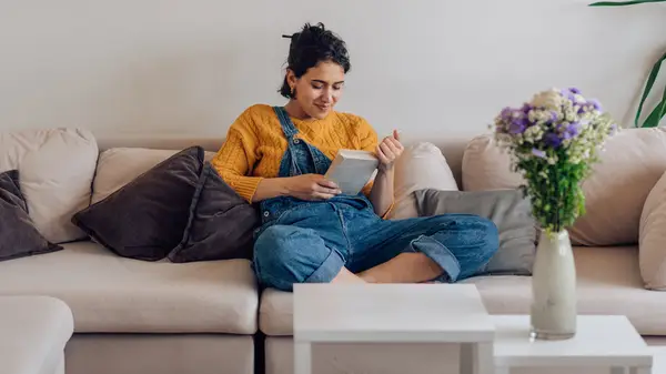 Brunette woman in denim overalls reading a book on a biege couch with a small smile