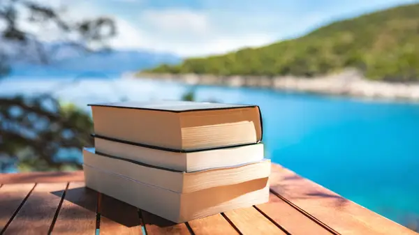 Books stacked on a wooden table with a picturesque body of water in the background