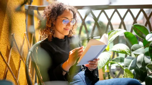 Curly-haired woman with glasses reading a book on a sunny balcony
