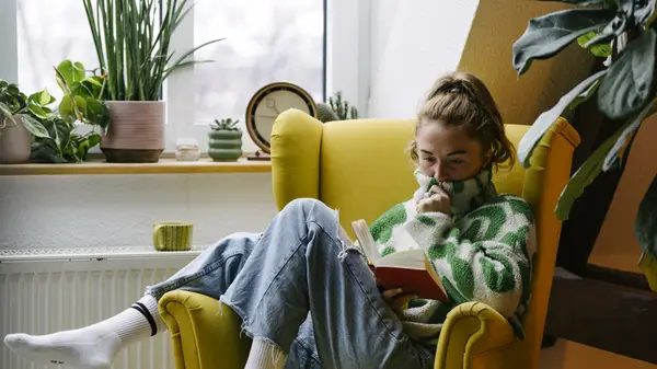 A woman sitting in a yellow armchair and reading a book