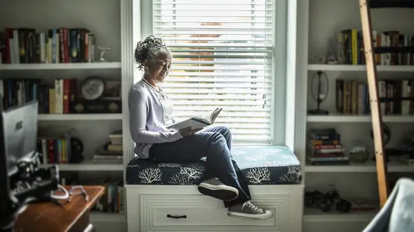 An older woman sitting in a window seat and reading a book