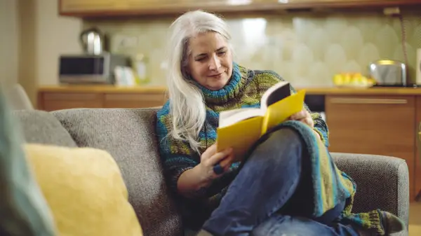 An older woman reads a book on the couch