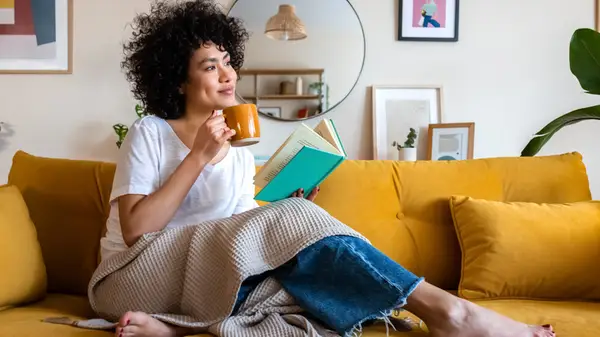 A woman holds a mug and book while sitting comfortably on the couch