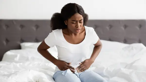 Woman sits on bed, gripping her lower abdomen in discomfort 